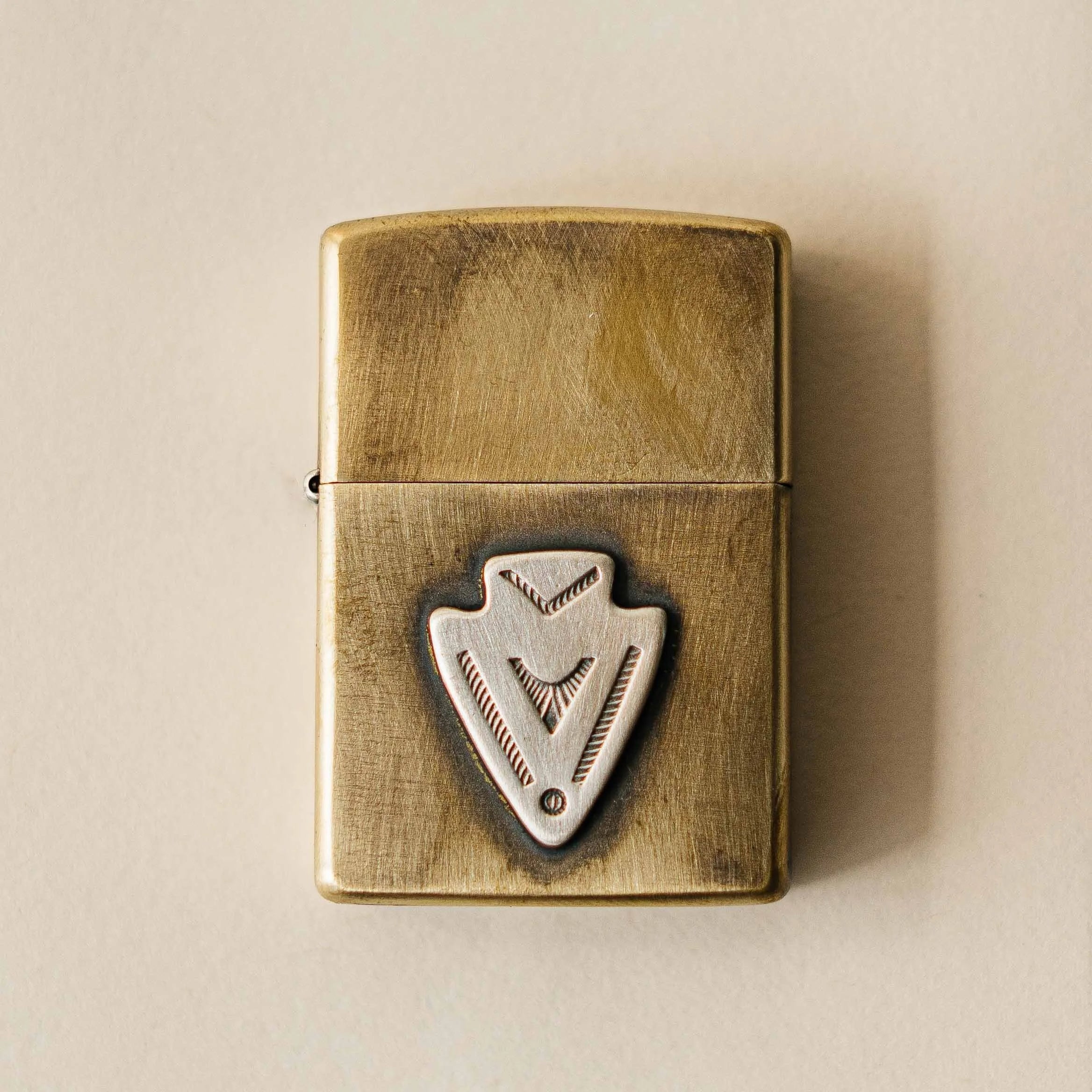 A brass Zippo lighter mounted with a sterling silver arrowhead, hand-finished with a patina that deepens with every year of carry