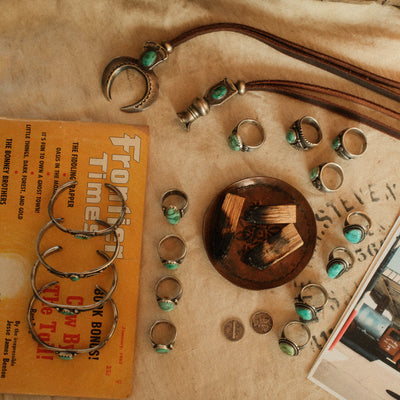 A styled flat-lay of multiple silver-toned rings with turquoise stone accents, a silver crescent moon pendant on a cord, and a silver-toned bracelet, arranged on a beige fabric surface with a yellow book, coins, and a small wooden dish.